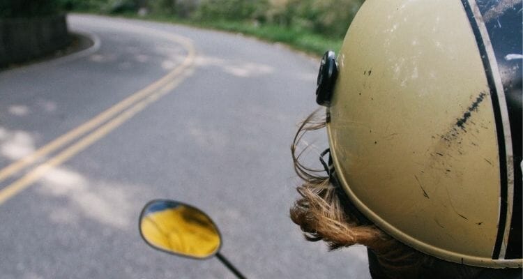 A motorcyclist walks down the street wearing an old helmet.